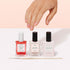 A hand with manicured nails hovers above three bottles of Manucurist French Manicure Glow polish and a thin brush on a white surface, highlighting gel-like shine against a light pink background.