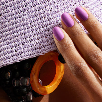 A hand with smooth Violette nail polish by Manucurist touches a lavender woven hat. Chunky orange earrings and black beaded accessories add stylish flair in the background.