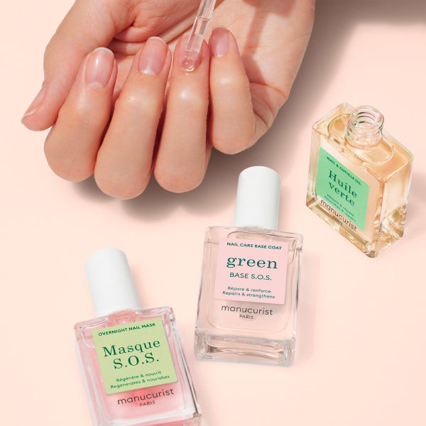 A hand has a dropper applying nail care oil to the nails. Three bottles of nail treatment products labeled Masque S.O.S., Green Base S.O.S., and Huile Verte are arranged on a light pink surface.