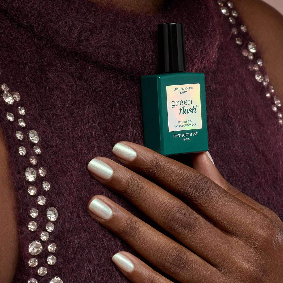 A hand with pearly white gel polish rests on a purple textured top with shiny rhinestones, holding a teal bottle of Fairy nail polish by Manucurist for ultimate sparkle nails.