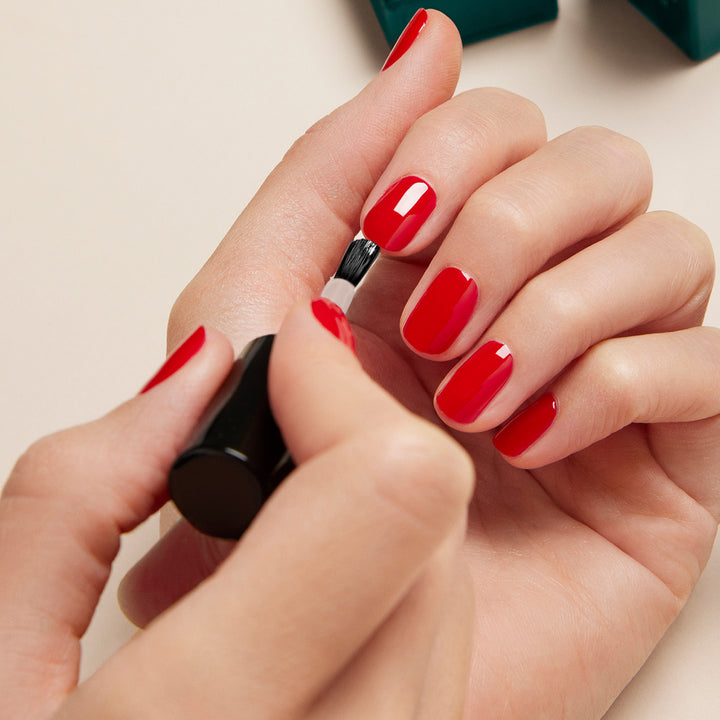 A close-up of a person applying a glossy red nail polish to their fingernails on one hand, while holding the nail polish brush in the other hand. The background is neutral and minimalistic.