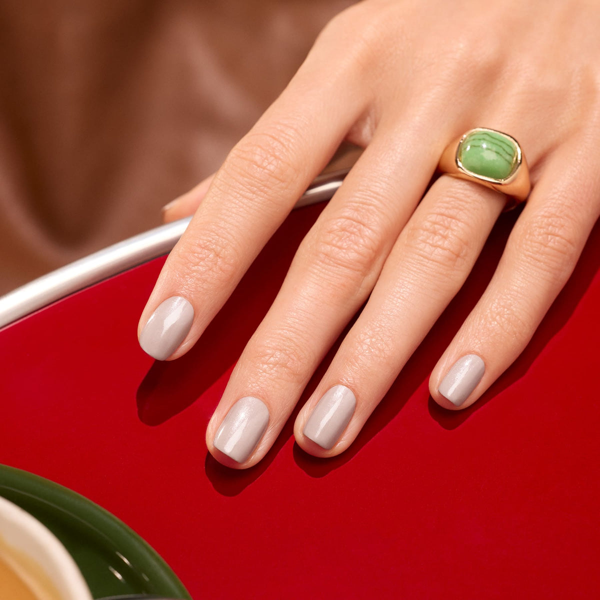 Une main aux ongles soigneusement manucurés, peints dans la teinte Tweed de Manucurist, un gris doux aux sous-tons nacrés, repose sur une surface rouge brillante. La personne porte à l’index une imposante bague en or sertie d’une grande pierre verte.