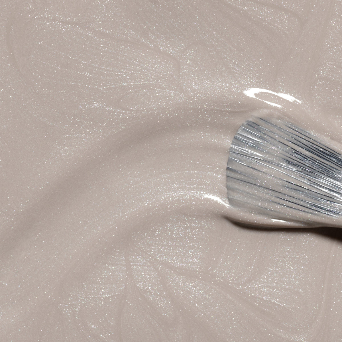 Gros plan d’un pinceau appliquant Manucurist Tweed, un vernis à ongles beige clair scintillant aux sous-tons nacrés, créant de délicats tourbillons dans la finition brillante.