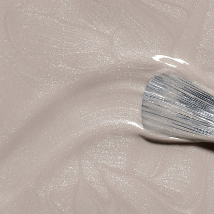 Gros plan d’un pinceau appliquant Manucurist Tweed, un vernis à ongles beige clair scintillant aux sous-tons nacrés, créant de délicats tourbillons dans la finition brillante.