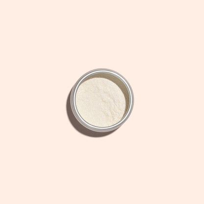 Une petite boîte métallique de Golden Glazed Effect Powder de Manucurist — une fine poudre blanc cassé qui donne aux ongles un scintillement doré nacré — est centrée sur un fond beige clair et projette une douce ombre vers la gauche.