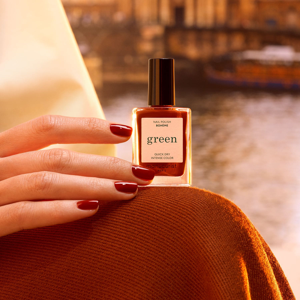 Une main portant le vernis rouge foncé aux sous-tons bruns Bohème de Manucurist, inspiré de Montmartre, tient un flacon de vernis vert sur un arrière-plan extérieur chaud et flouté pour une ambiance chaleureuse et élégante.