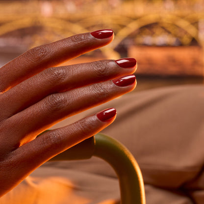 Gros plan sur une main à la peau brun foncé aux ongles soigneusement manucurés vernis avec Bohème, un vernis rouge brillant signé Manucurist, sur un fond chaud doucement flouté.
