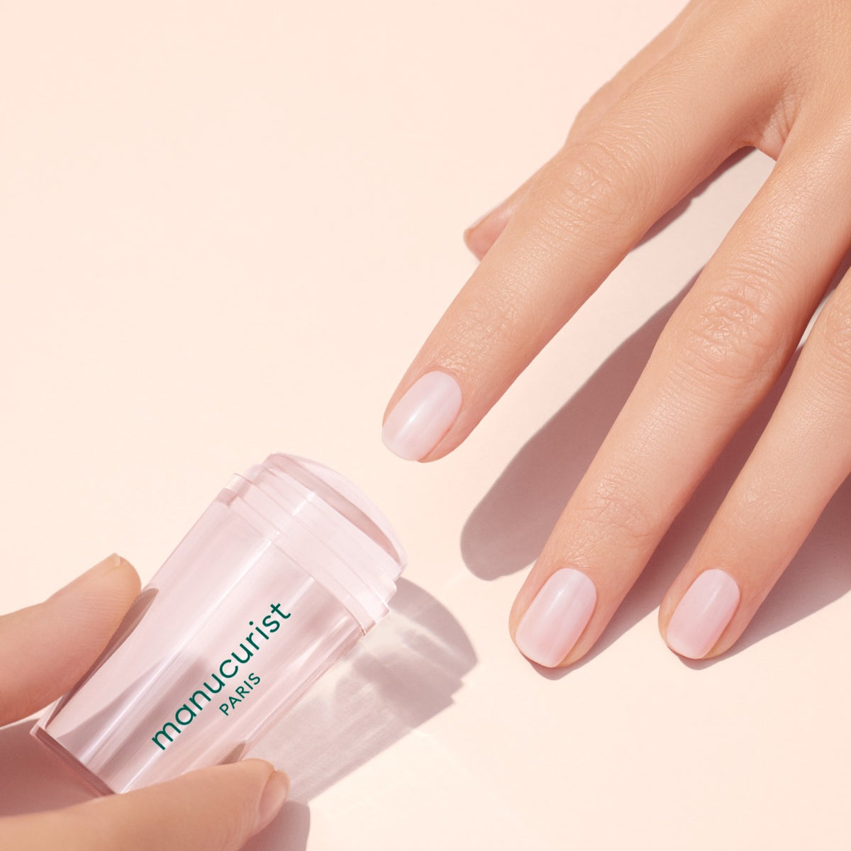 A hand with neatly manicured, natural nails touches a soft pink surface. Another hand holds a pink manicure product labeled manucurist PARIS. The scene is bright and minimalistic.