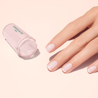 A hand with neatly manicured, flawless French tips rests next to a translucent pink Mani stamper by Manucurist US on a light pink surface.