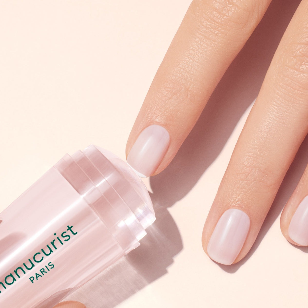 A hand with neatly manicured, natural-looking nails is shown using a clear, cylindrical nail care product labeled manucurist PARIS on a light pink background.