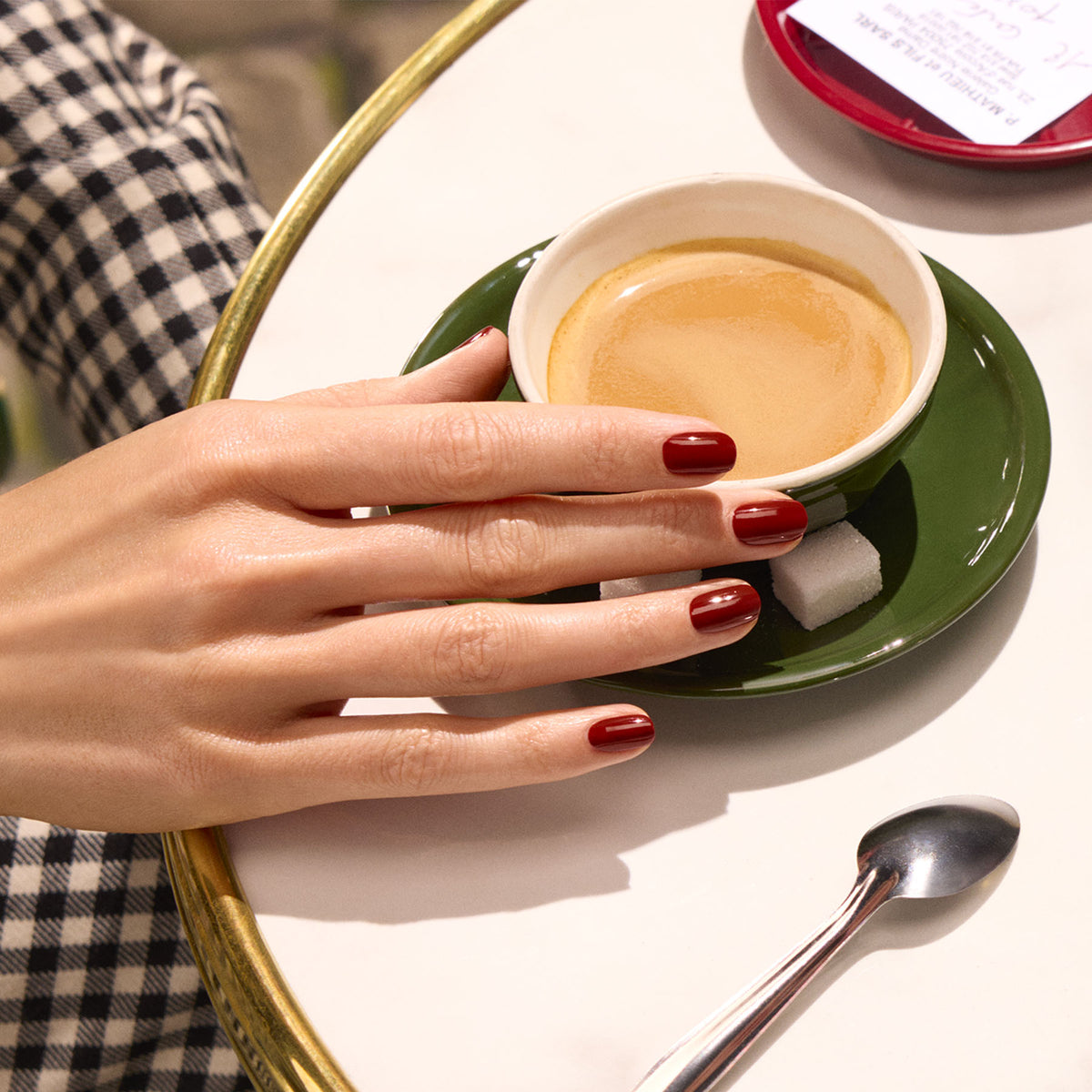 Une main avec le vernis rouge brillant Bohème de Manucurist repose sur une soucoupe accueillant une tasse d&