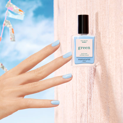 A neatly manicured hand with pastel blue-grey nails in the shade "Bubble" by Manucurist is shown beside its matching bottle, set against a pale pink wooden backdrop and a bright blue sky with colorful bunting.