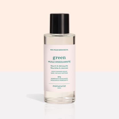 A clear bottle with a black cap contains Manucurist Nail Polish Removing Oil, a green, plant-based formula. The bottle features a light pink label with green text on a beige and white background.