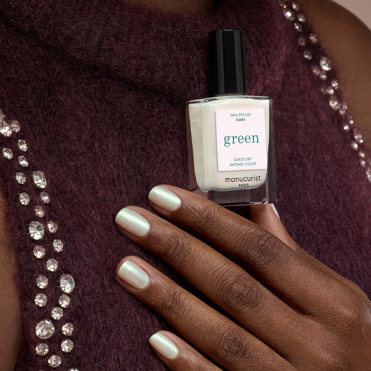 A hand with light green metallic sparkle nail polish holds a matching bottle labeled "Fairy" by Manucurist, set against a maroon top adorned with rhinestones.