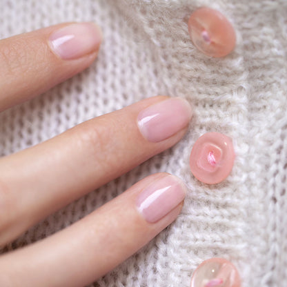 Une main aux ongles soigneusement manucurés, peints en Hortencia de Manucurist, un vernis rose transparent, repose sur un doux tissu blanc tricoté décoré de boutons roses.
