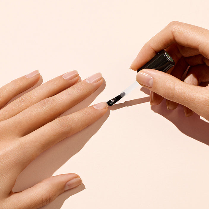 A person applies clear nail polish to their fingernails on one hand using a brush, while the other hand rests on a light beige surface.