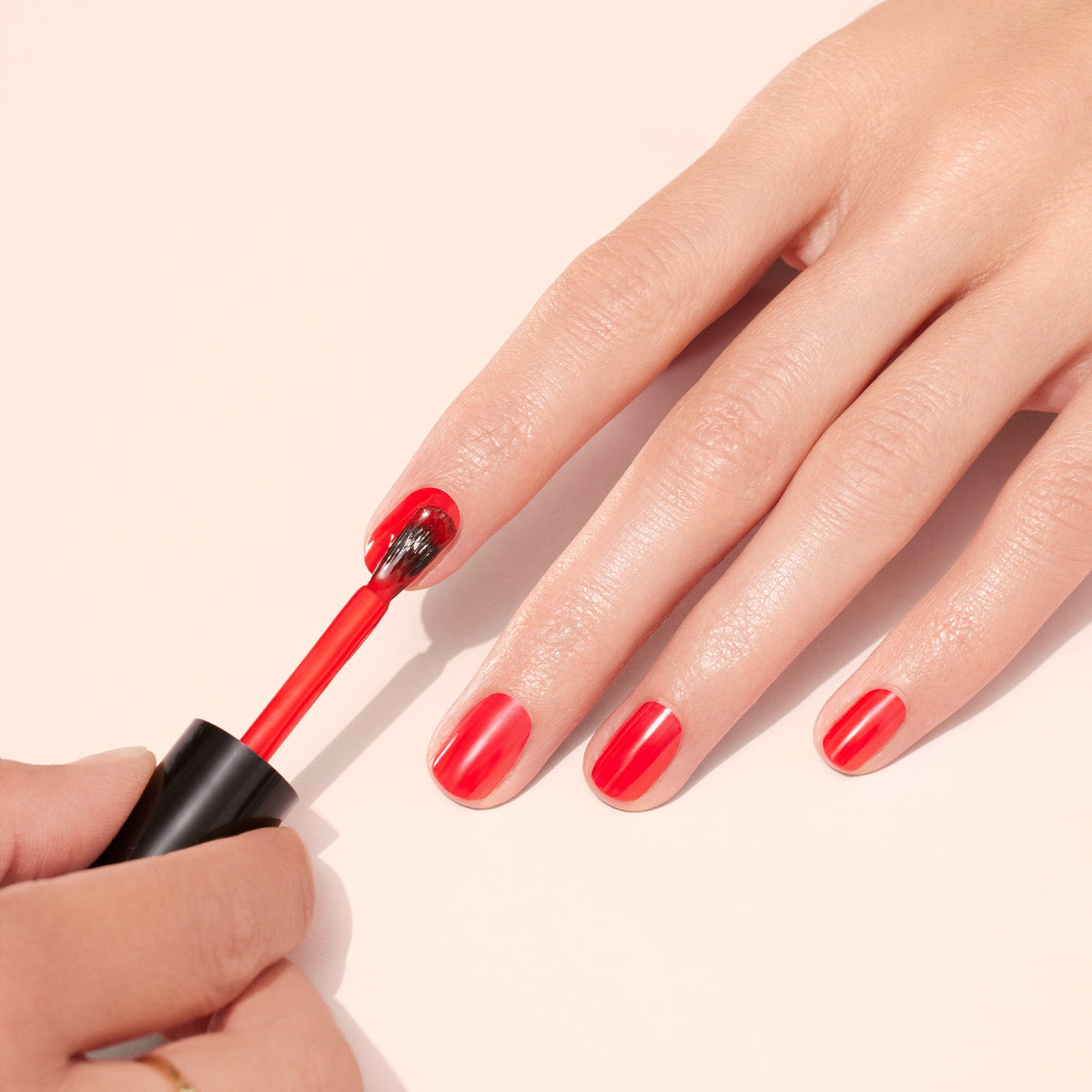 A hand is having its fingernails painted with bright red nail polish. Another hand holds the brush, applying polish to the fingernails on a light, neutral background.