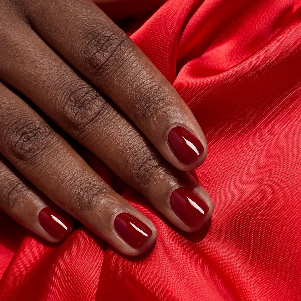 A close-up of a hand with neatly manicured nails painted in Dark Pansy by Manucurist, resting on vibrant red satin fabric.