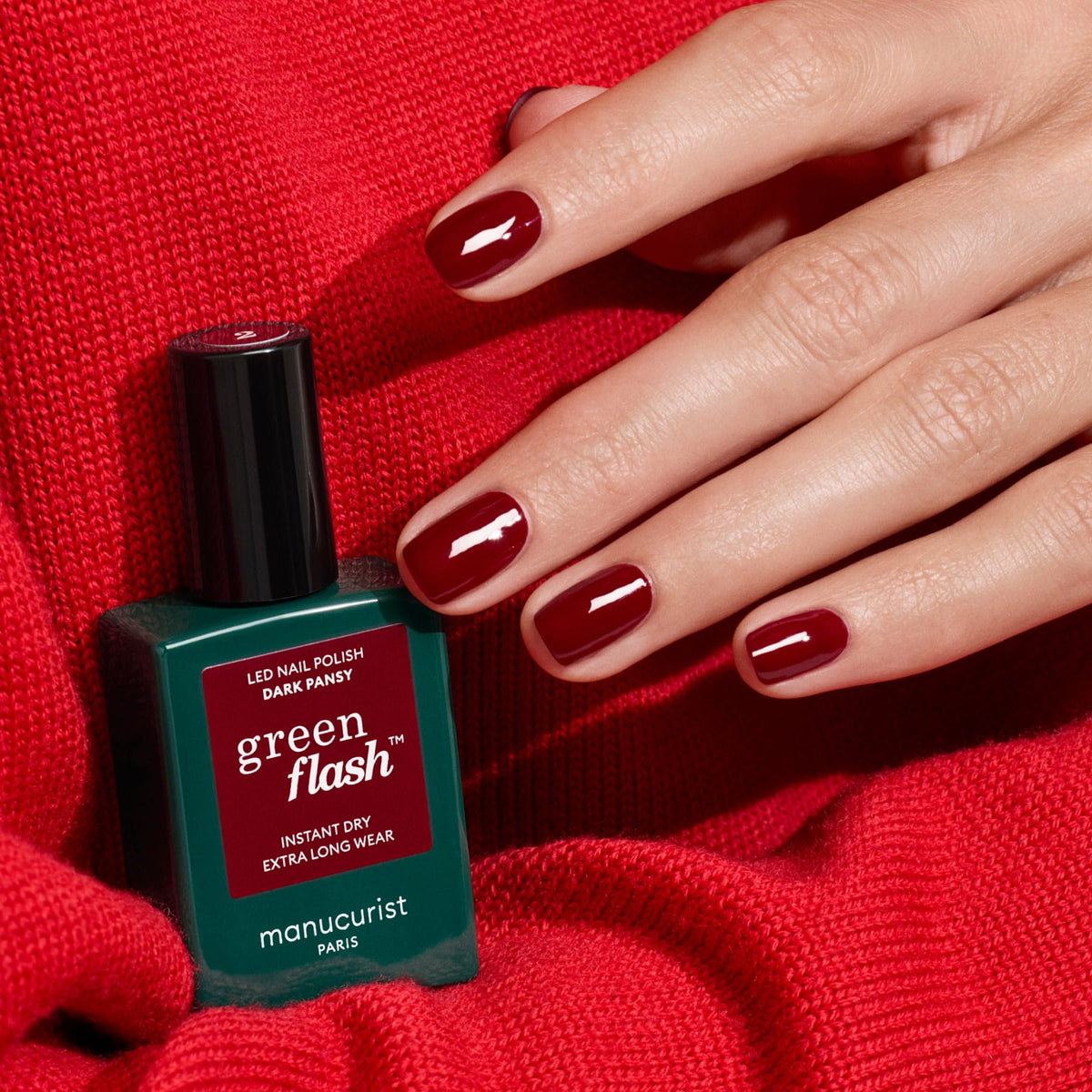 A hand with deep red, perfectly manicured nails rests on red knit fabric beside a Manucurist bottle of Green Flash gel polish in the Dark Pansy shade.