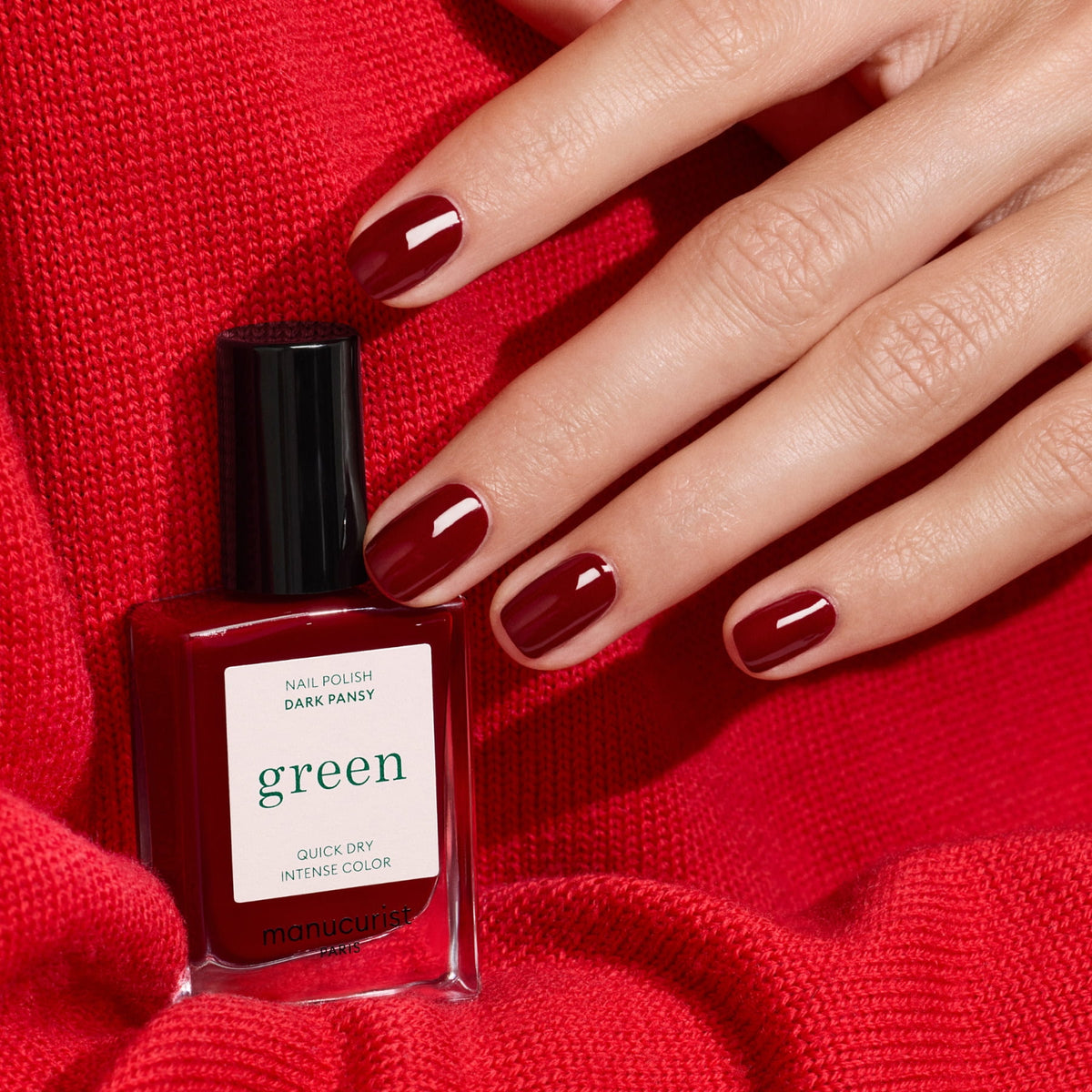 A hand with short, neatly manicured nails painted velvet red rests on red fabric beside a matching bottle of Manucurist nail polish in the shade Dark Pansy.