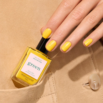 Une main aux ongles soigneusement manucurés, vernis en Gold Button par Manucurist, tient un flacon de vernis à ongles Gold Button jaune assorti, posé sur un fond de tissu beige.