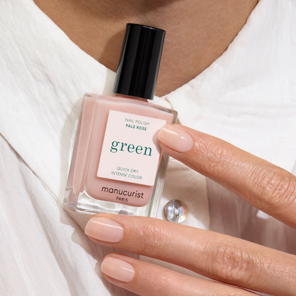 Une main aux ongles soigneusement manucurés tient un flacon de vernis à ongles Manucurist Pale Rose, avec une chemise blanche visible en arrière-plan.