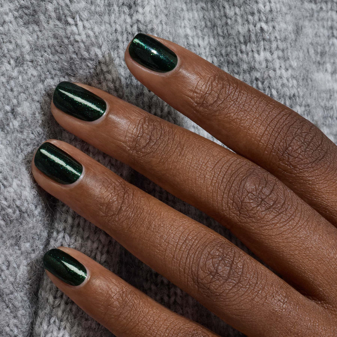 A close-up of a hand with glossy Manucurist Poison gel polish in forest green, resting on light gray knit fabric.