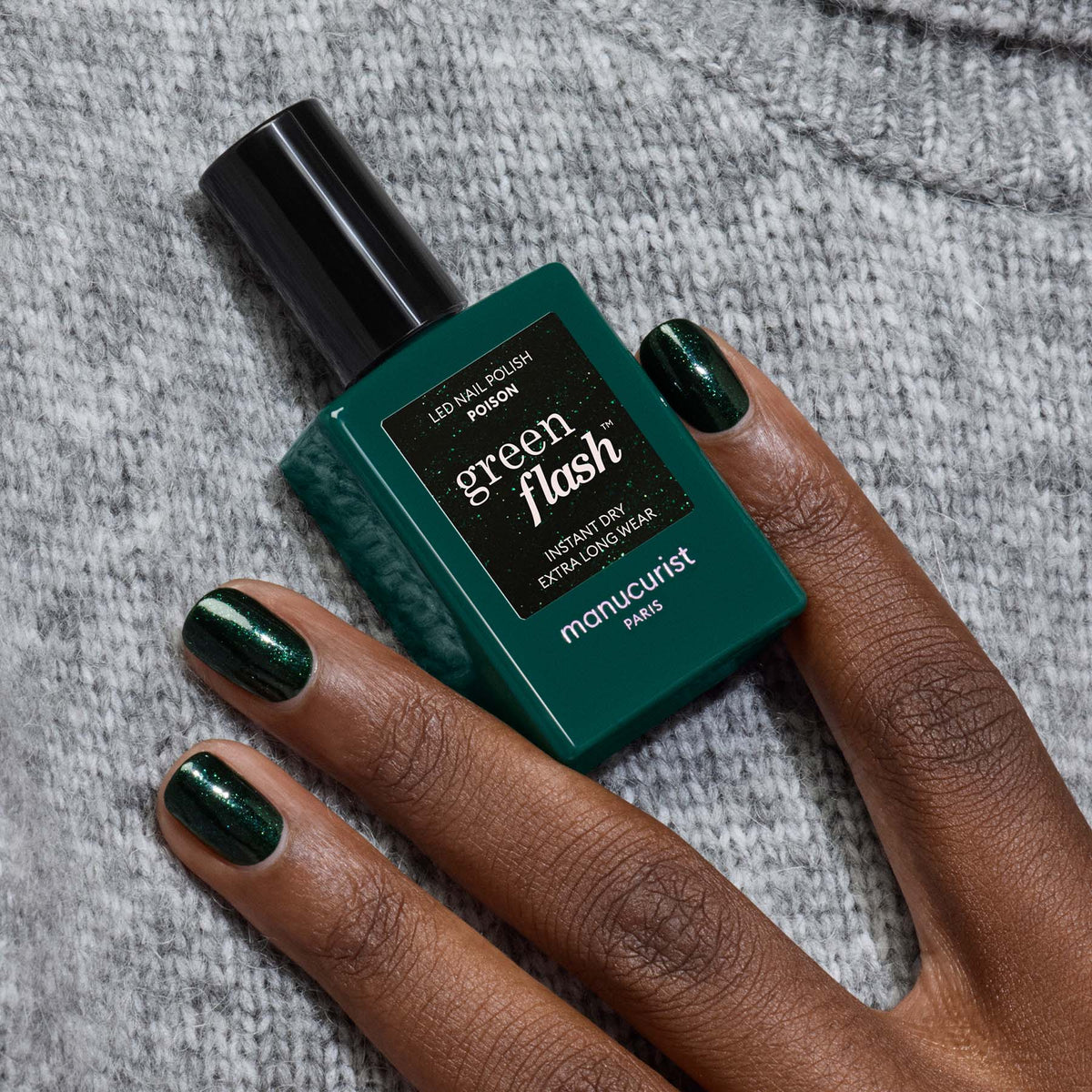 A hand with dark, glossy forest green gel polish holds a matching bottle of Manucurist “Poison” nail polish against a gray knitted fabric background.