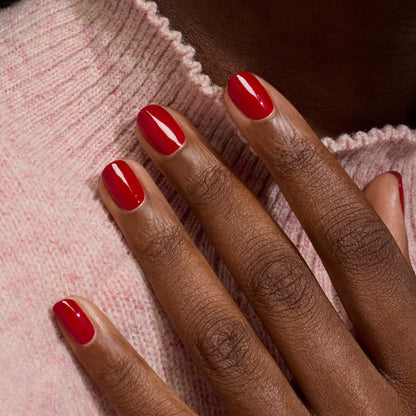 Gros plan sur une main à la peau brun foncé et aux ongles manucurés vernis avec le vernis gel Red Velvet de Manucurist, posée sur un pull en maille rose clair.