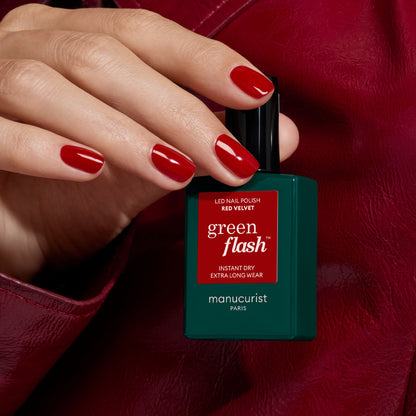 Une main aux ongles vernis de rouge tient un flacon de vernis Manucurist Red Velvet sur un fond en cuir rouge.