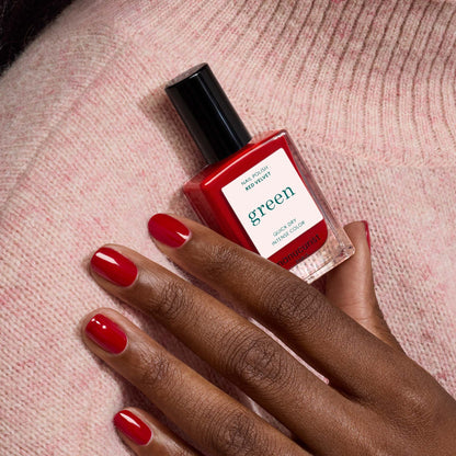 A hand with neatly manicured velvet red nails holds a square bottle of Manucurist Red Velvet nail polish from the Green range against a soft pink knitted fabric background.
