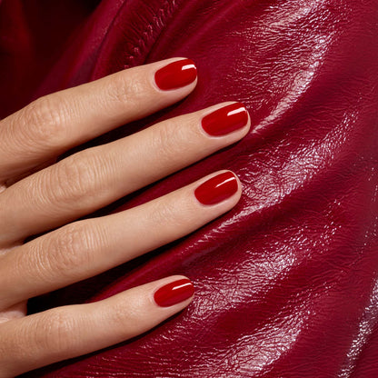 Gros plan sur une main aux ongles impeccablement manucurés avec le vernis Red Velvet de Manucurist, posée sur une surface rouge brillante et texturée.