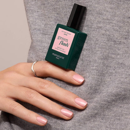 A hand with pale pink nail polish holds a bottle of Manucurist Satin nail polish against a gray sweater.