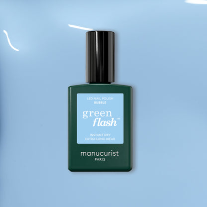 A bottle of Manucurist Bubble nail polish with grey undertones rests on a smooth light blue surface. The dark green bottle features a light blue label and black cap.