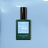 A bottle of Manucurist Bubble nail polish with grey undertones rests on a smooth light blue surface. The dark green bottle features a light blue label and black cap.