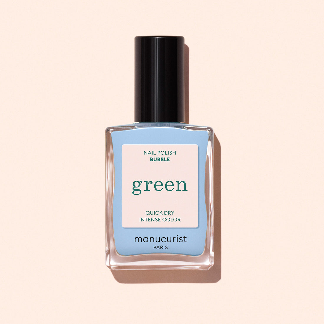 A bottle of Manucurist Bubble nail polish rests on a light beige background. It features a black cap and a pastel blue and cream label with green and pink text, suggesting light blue polish with grey undertones.
