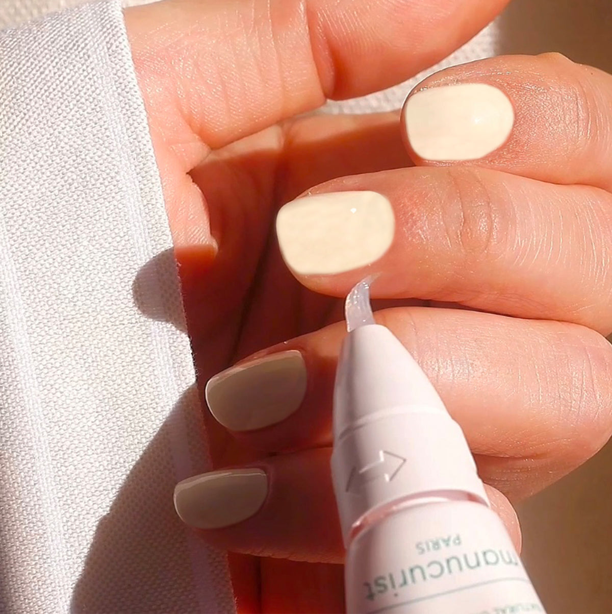 A hand with short, neatly manicured nails painted in a pale, creamy polish holds a white pen-like applicator close to the nails, likely for nail care or cuticle treatment. Sunlight gently illuminates the fingers.