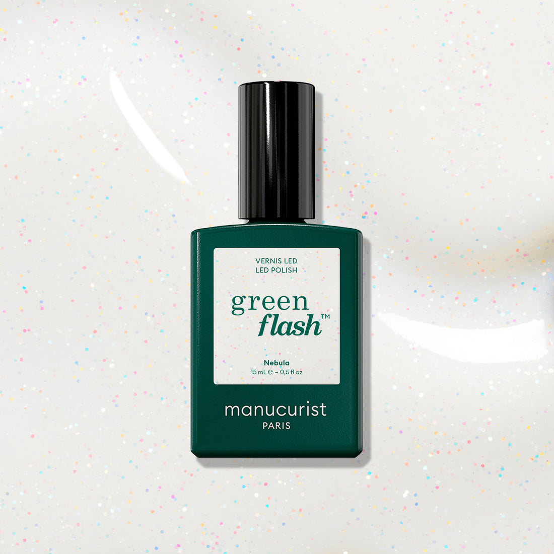 A bottle of Manucurist Nebula nail polish rests on a white surface with colorful specks. The green bottle, labeled Nebula, suggests a holographic effect under its sleek black cap.