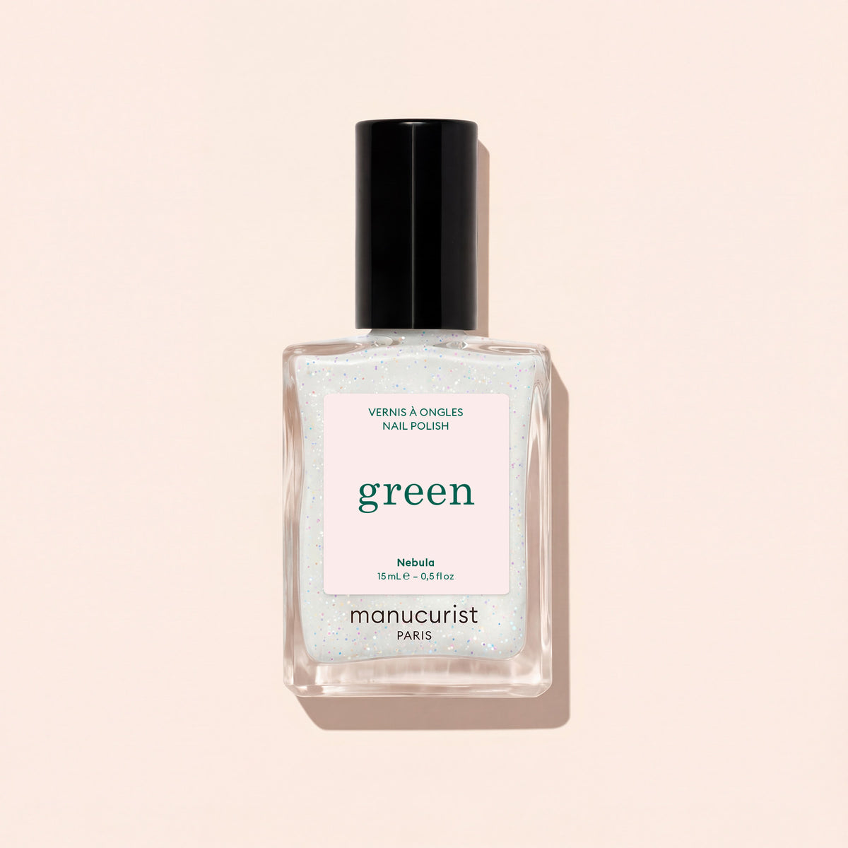 A clear square bottle of Manucurist Nebula nail polish with a black cap and white label, filled with shimmery holographic polish, is photographed on a pale beige background.
