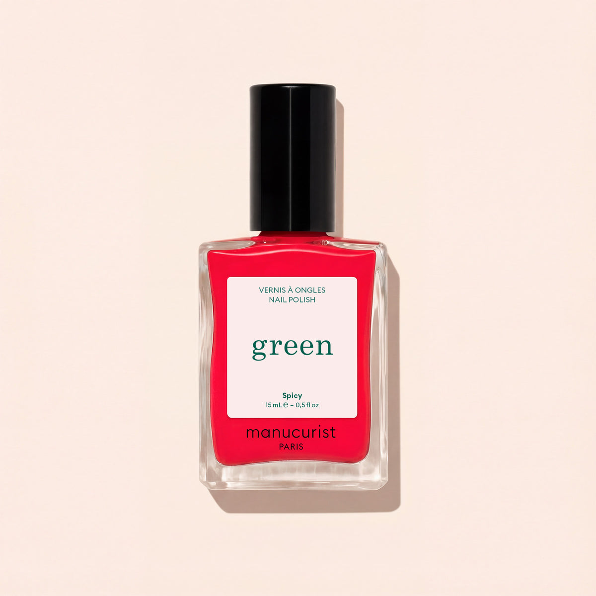 A bottle of Manucurist Spicy nail polish, a pop red shade with pink undertones from the Green™ range, features a black cap and a white label, set against a light beige background.