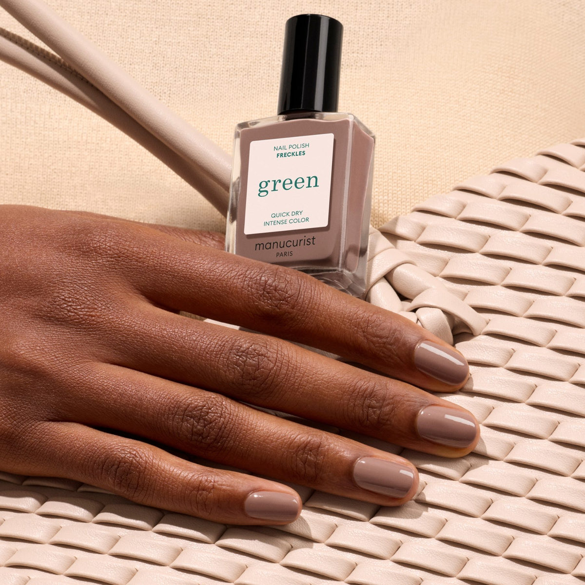 A hand with neatly manicured Freckles by Manucurist nails rests on a woven cream bag, holding the polish bottle to showcase chic style against a beige background.