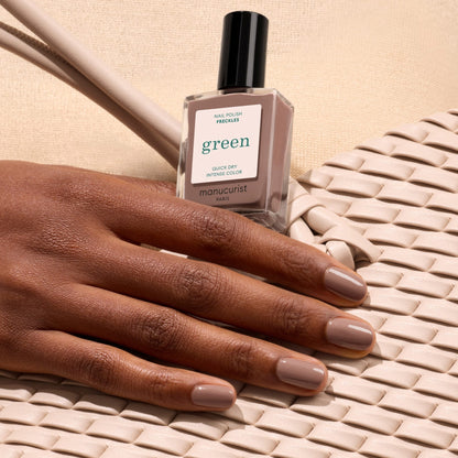 A hand with neatly manicured Freckles by Manucurist nails rests on a woven cream bag, holding the polish bottle to showcase chic style against a beige background.