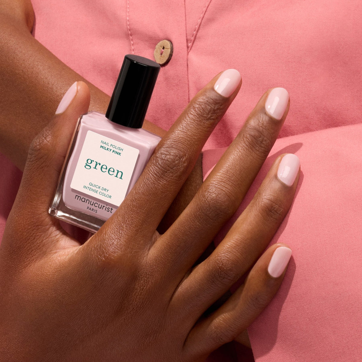 A hand with neatly manicured Milky Pink nails from Manucurist holds the bottle against a pink shirt background.