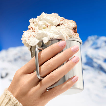 A hand with Manucurist’s Milky Pink nail polish holds a metal mug of hot chocolate topped with whipped cream, set against snowy mountains and a bright blue sky.