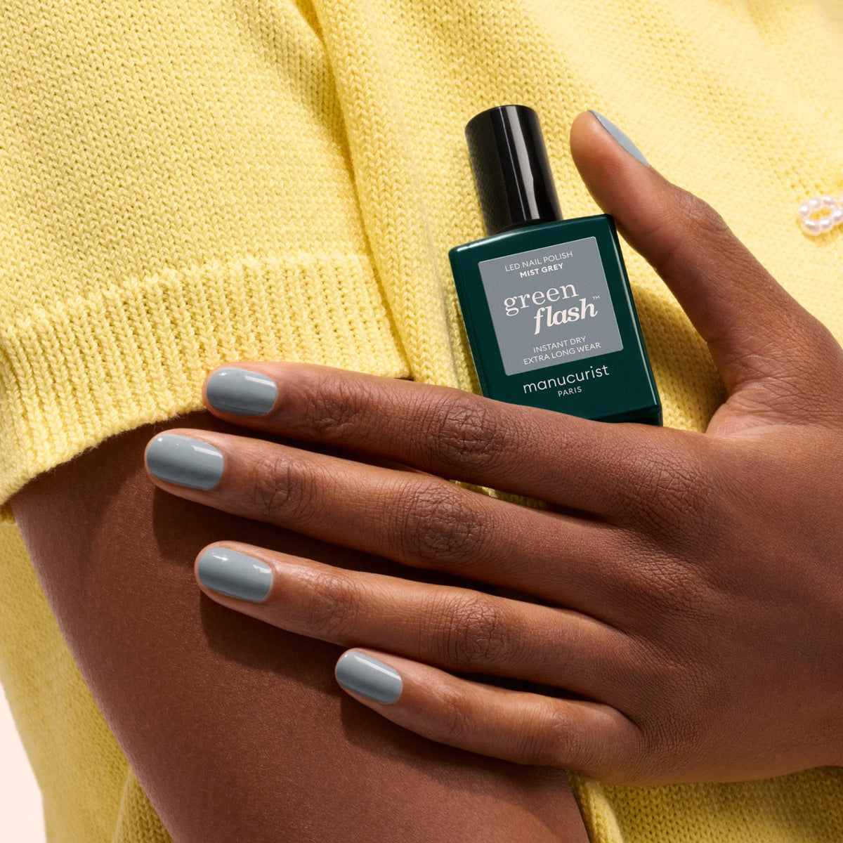 Une personne à la peau mate met en valeur ses ongles vernis en gel Mist Grey tout en tenant un flacon de vernis Manucurist sur un haut en maille jaune.