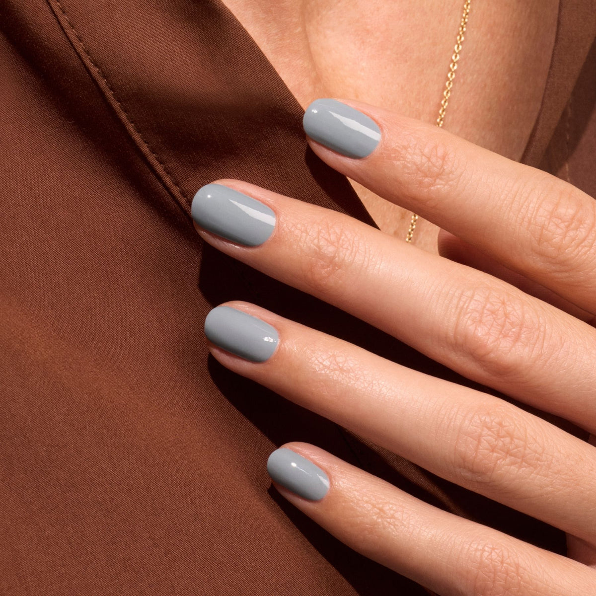 A close-up of a hand with short, neatly manicured nails painted Mist Grey by Manucurist rests on brown fabric, with a delicate gold necklace partially visible in the background.