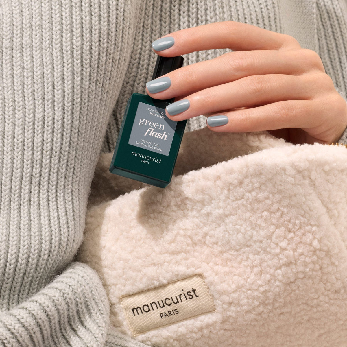 Une main aux ongles peints en Mist Grey tient un flacon de vernis gel Green Flash de Manucurist, portant un pull en maille grise et tenant un article sherpa crème avec une étiquette manucurist PARIS.