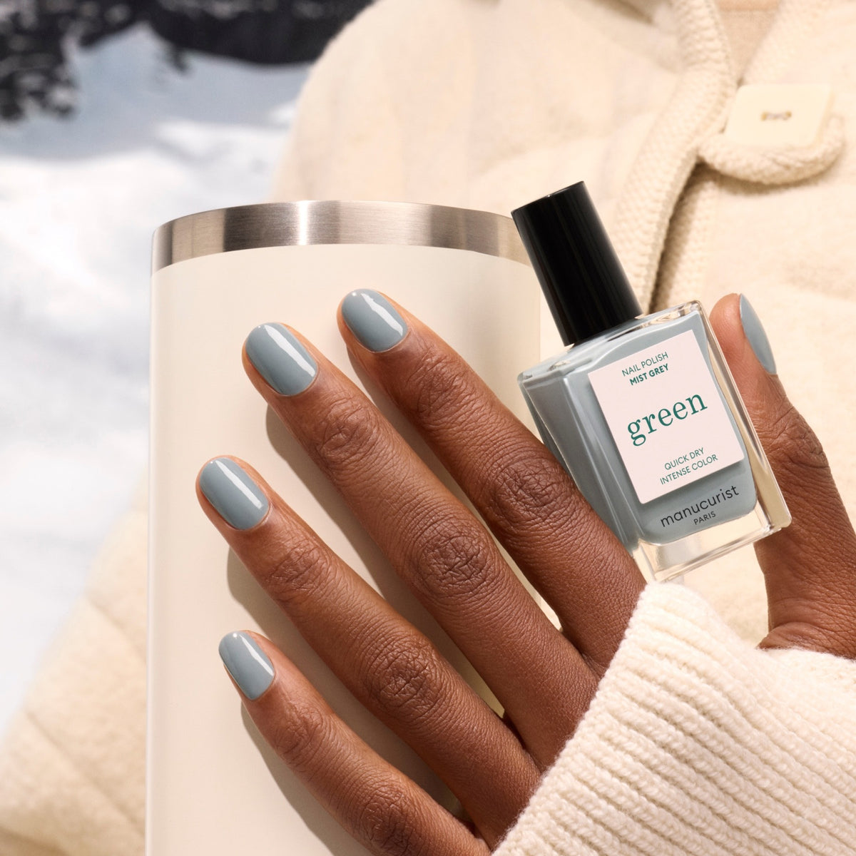 A hand with muted grey painted nails holds a white tumbler and a bottle of Manucurist Mist Grey nail polish in the same shade while wearing a cream-colored coat.