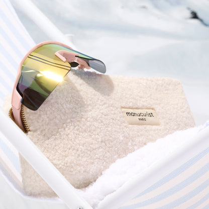 A pair of reflective sunglasses sits on the soft Teddy Bag by Manucurist-us, placed on a white and blue striped surface with a snowy backdrop—a chic winter accessory.