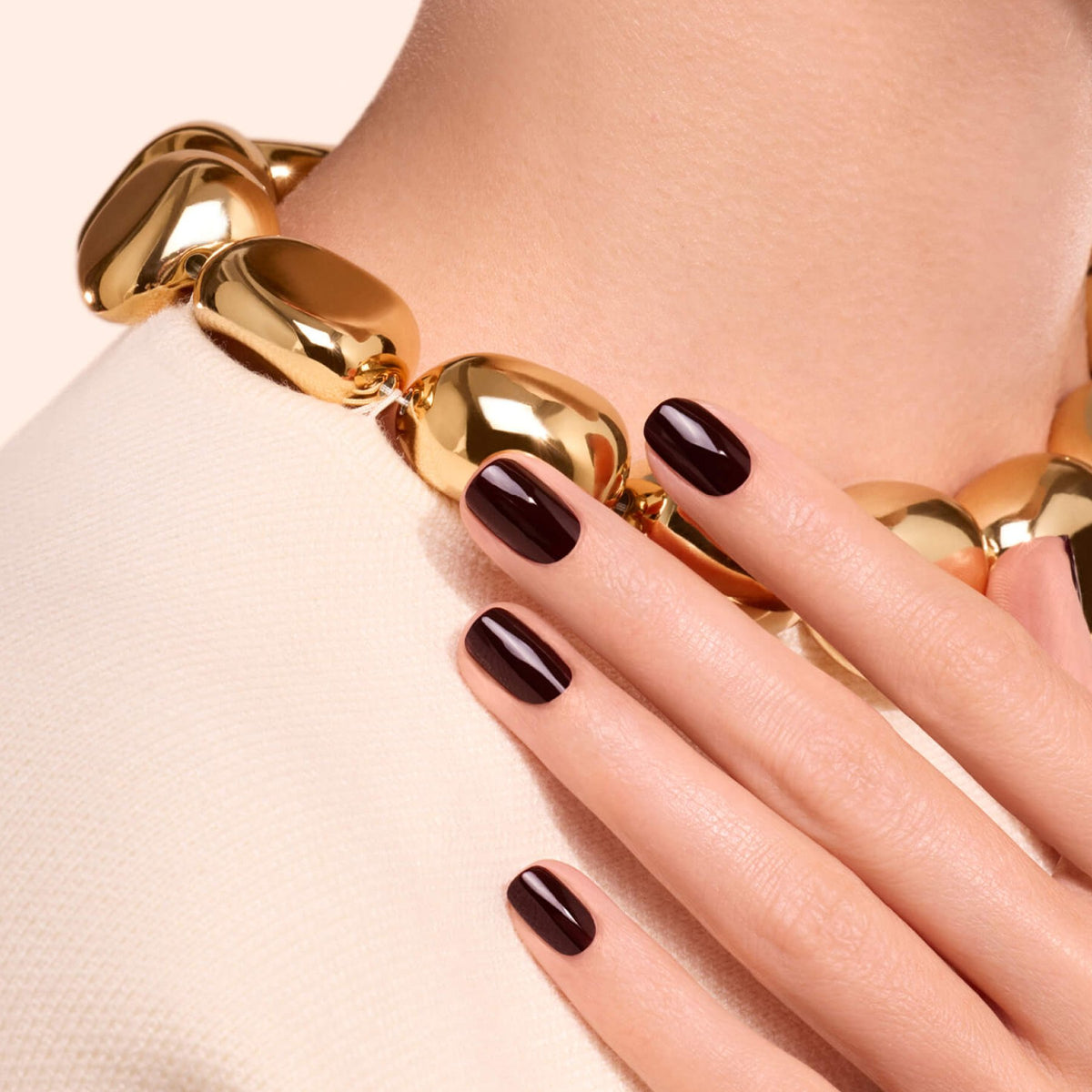 A hand adorned with Manucurist’s Hollyhock—a deep red nail polish—rests on a textured white top. The person wears a chunky gold chain necklace, set against a pale, simple background.